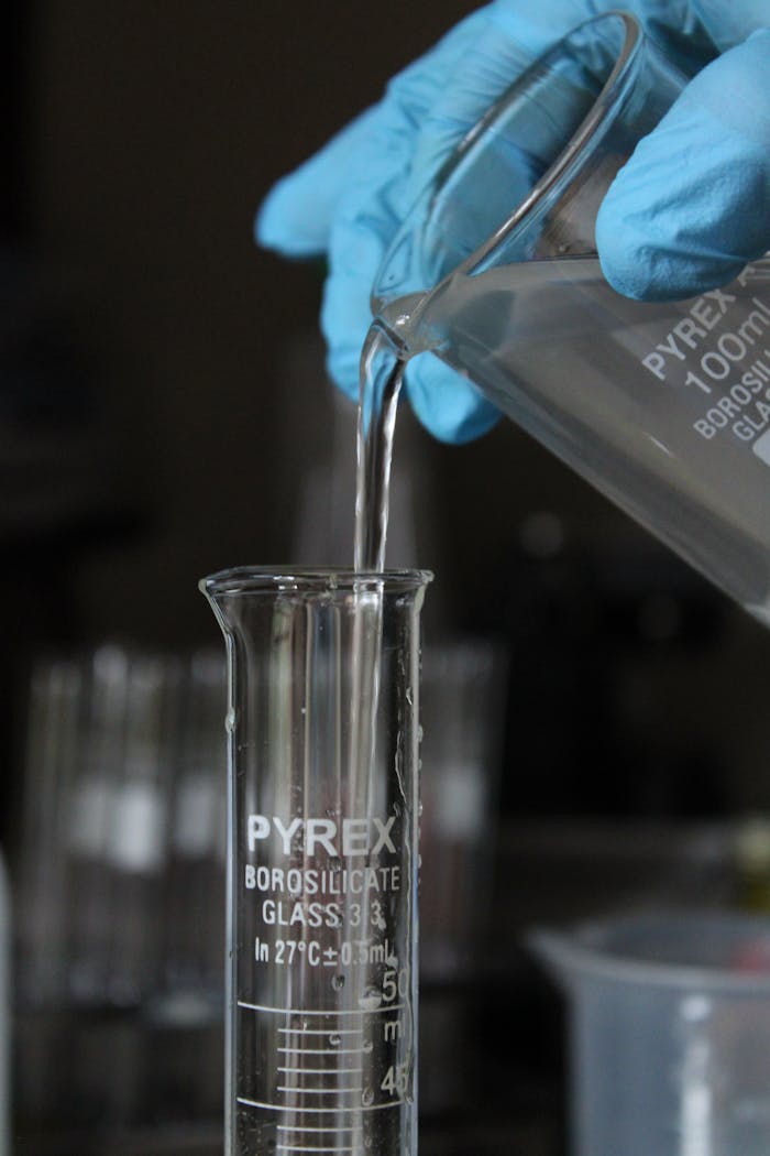 Scientist wearing gloves pours liquid into a measuring beaker in a laboratory setting.